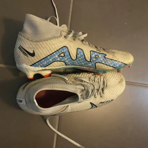 Nike Air Zoom fotbollsskor - Säljer ett par vita Nike Air Zoom fotbollsskor i storlek herr 41. Skorna har en hög ankelkrage och snörning för bra passform. De har en blå Nike-logga och texten 'AIR' på sidan. Skorna är i syntetmaterial och är i välanvänt skick. Perfekta för fotbollsträning och matcher. De är ganska små i storleken, jag har vanligtvis 39 i skor.