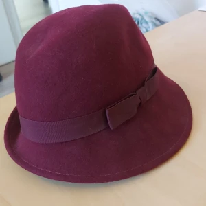 Vinröd hatt med rosett - Säljer en snygg vinröd hatt med en matchande rosett på sidan. Hatten är gjord av ett mjukt material och har en klassisk form som passar perfekt för höst och vinter. Den är i dam one size från Åhléns och i nyskick. Perfekt för att ge din outfit en elegant touch!