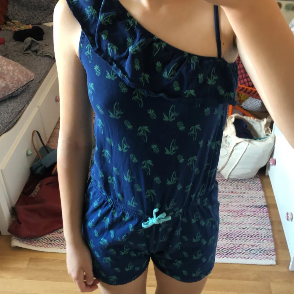 Säljer en superfin mörkblå oneshoulder jumpsuit från Pepperts! med ett tropiskt mönster av palmer och ananaser. Jumpsuiten har en volangdetalj vid axeln och ett knytband i midjan för en bekväm passform. Perfekt för sommarens alla fester och äventyr! 🌴🍍. Mekot.