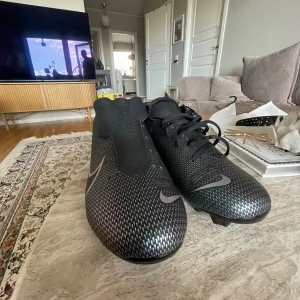 Svarta fotbollsskor från Nike - Säljer ett par svarta fotbollsskor från Nike. Skorna har en snygg design med små prickar och en diskret rosa Nike-logga på sidan. De har snörning och är i nyskick. Perfekta för fotbollsträning eller match!