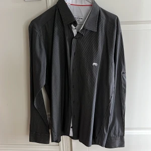 Svart och vit randig skjorta från Moods of Norway - Säljer en snygg svart och vit randig skjorta från Moods of Norway. Skjortan är i storlek L och är tillverkad av 100% bomull. Den har långa ärmar och knappar framtill. Perfekt för både vardag och fest!