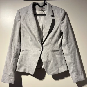 Grå kavaj - Säljer en stilren och klassisk grå kavaj med långärmad design. Perfekt för både kontoret och mer formella tillfällen. Kavajen har en enkel knäppning framtill och är figursydd för en snygg passform.