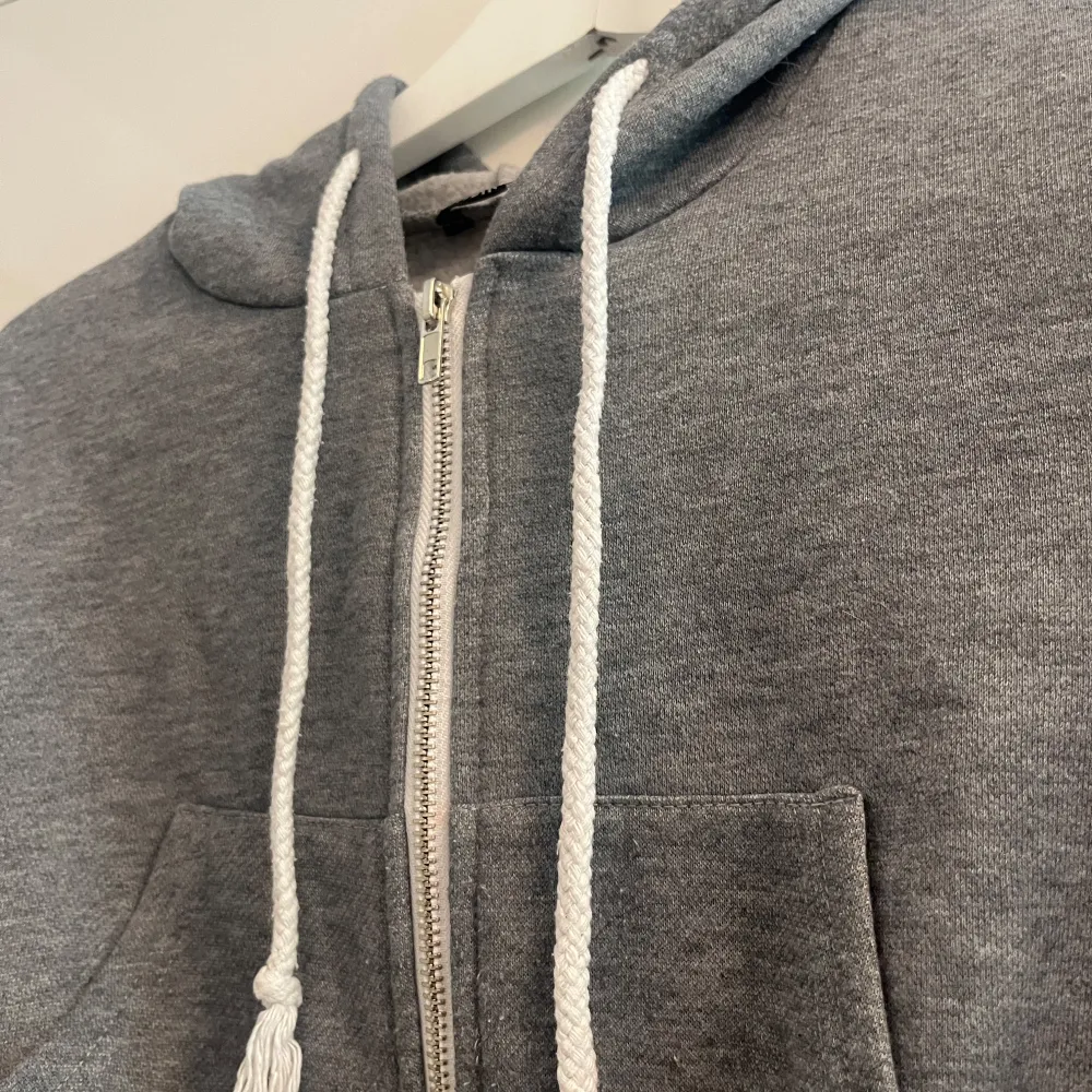 Säljer en supermysig grå croppad hoodie från Shein. Den har en dragkedja framtill och en stor luva med vita snören. Perfekt för en chill dag eller när du vill ha en avslappnad stil. Den är i storlek XS och passar perfekt till höst och vår.. Tröjor & Koftor.