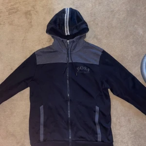 Svart och grå huvtröja från Boss - Säljer en snygg svart och grå huvtröja från Boss. Den har en dragkedja framtill och en huva med snörning. Perfekt för en avslappnad stil!