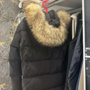 Svart dunjacka med pälskrage - Varm svart jacka med päls från hollies. Jackan har använts under två vintrar och köptes  för 6000kr.