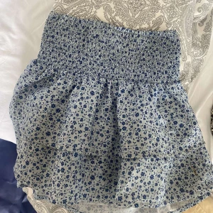 Blommig kjol från Gina Tricot - Säljer en superfin blommig kjol från Gina Tricot. Den är blå och vit med  sött blommönster. Kjolen har en smockad midja som ger en bekväm passform och volanger som ger en extra touch av stil. Perfekt för sommardagar eller en avslappnad vårutflykt!