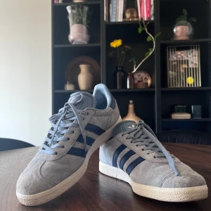 Blå Adidas Gazelle sneakers - Säljer ett par snygga ljusblåa Adidas Gazelle sneakers i mocka. De har de klassiska tre ränderna i mörkblått på sidan och en vit sula. Skorna är i bra skick och sparsamt använda. Säljes endast för att jag har för många skor. 