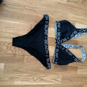 Svart bikini med textdetaljer - Säljer en snygg svart bikini med textdetaljer på banden där det står 'SURF + BEACH'. Bikinin har en sportig stil med breda axelband och en bekväm passform. Perfekt för en dag på stranden eller vid poolen!