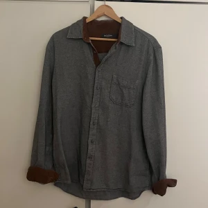 Grå skjorta med bruna detaljer - Snygg skjorta köpt begagnat men i bra skick, förutom att en av knapparna verkar ha sytts om vilket syns på bild 3 men knappt i verkligheten. Färgen på skjortan visas rättvisast på bild 1. Jag är 188 cm. Hör av dig vid minsta fråga eller fler bilder!