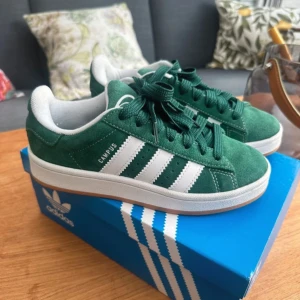 Gröna Adidas Campus sneakers - Säljer ett par snygga gröna Adidas Campus 00s sneakers i mocka. De har de klassiska vita ränderna på sidan och en vit sula. Skorna är i nyskick och kommer i originalkartong. Perfekta för både vardag och fest!