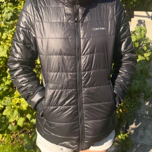 Svart pufferjacka från Calvin Klein - Säljer en snygg svart jacka från Calvin Klein. Jackan är lätt och har en quiltad design med dragkedja framtill. Den är perfekt för höst och vår, och har Calvin Klein-logga vid ärmsluten. Jackan är i nyskick och använd ett fåtal gånger. Storlek S skulle uppskatta att även skulle passa någon som har xs.