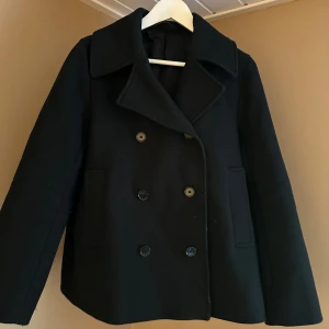 Svart skepparkavaj från COS - Säljer en stilren svart skepparkavaj från COS. Den har en klassisk dubbelknäppt design med stora knappar och en bred krage. Perfekt för höst och vinter, och ger en elegant touch till vilken outfit som helst. Jackan är i bra skick och redo för nya äventyr!