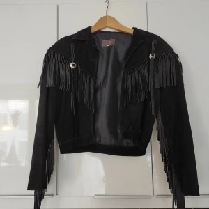 Svart mockajacka med fransar från Svelte Leather - Säljer en supercool svart mockajacka från Svelte Leather, Melbourne. Köpt i Australien. Den har långa ärmar och är dekorerad med fransar både fram och bak, vilket ger den en unik och edgy look. 