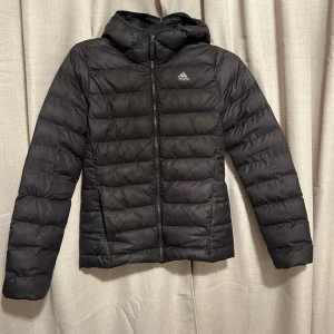 Svart dunjacka från Adidas - Säljer en svart dunjacka från Adidas i storlek XS. Jackan har en dragkedja framtill och en huva med tre ränder. Perfekt för kyliga dagar med sin quiltade design som håller värmen. Den är i nyskick och redo för höst- och vinteräventyr!