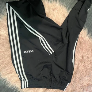 Svarta träningsbyxor från Adidas - Säljer ett par svarta träningsbyxor från Adidas med de klassiska tre vita ränderna längs sidorna. Byxorna har en elastisk midja och är perfekta för träning eller en avslappnad dag. De finns mindre defekter 