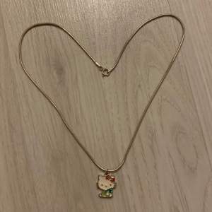 silvrigt halsband med hello kitty på.  hon har ljusgröna kläder och håller i ett äpple i mörkare grön färg, med sin ikoniska roset i rödrosa. bra kvalité och jättegulligt!! kontakta mig för fler bilder eller frågor. 