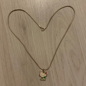 Hello Kitty halsband - silvrigt halsband med hello kitty på.  hon har ljusgröna kläder och håller i ett äpple i mörkare grön färg, med sin ikoniska roset i rödrosa. bra kvalité och jättegulligt!! kontakta mig för fler bilder eller frågor. 