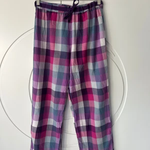 Rutig byxa i lila och rosa toner - Säljer ett par pyjamasbyxor från Åhléns i storlek 36/38 (S). De har ett snyggt rutigt mönster i lila, rosa och blå nyanser. Byxorna har släppt i sömmen vid snörningen och på baksidan. Men fortfarande användbara. Har noppror på sig.