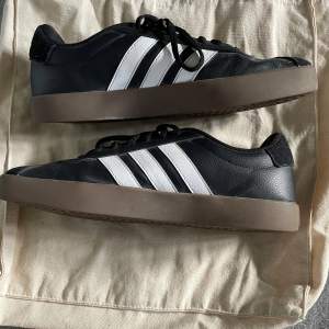 Säljer ett par snygga svarta Adidas sneakers med de klassiska tre vita ränderna på sidan. Skorna har en brun sula och är i bra skick. Perfekta för vardagsbruk och ger en stilren look. De har snörning för en bra passform.