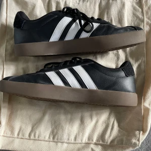Svarta Adidas sneakers med vita ränder - Säljer ett par snygga svarta Adidas sneakers med de klassiska tre vita ränderna på sidan. Skorna har en brun sula och är i bra skick. Perfekta för vardagsbruk och ger en stilren look. De har snörning för en bra passform.