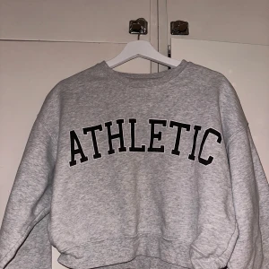 Grå sweatshirt från Bik Bok - Säljer en supermjuk och bekväm grå sweatshirt från Bik Bok i storlek XS. Den har texten 'ATHLETIC' tryckt i svart på framsidan. Perfekt för en avslappnad stil eller för att mysa hemma. Tröjan har en normal passform och långa ärmar.