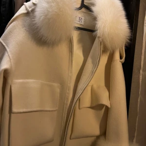 offwhite kappa med pälsdetaljer - Säljer en superfin offwhite kappa från fur Sthlm med fluffiga pälsdetaljer på kragen och armar, (går att ta bort) Den har en stilren design med stora fickor fram och en dragkedja. Perfekt för att hålla sig varm och snygg under höst och vinter!