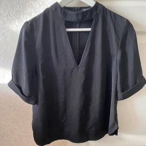 Svart blus med v-ringning - Säljer en stilren svart blus med v-ringning och trekvartsärmar. Blusen har en dragkedja i ryggen och är perfekt för både vardag och fest. Den är i bra skick och passar till många olika outfits.