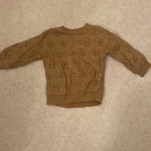 Brun tröja med Musse Pigg-mönster - Säljer en supergullig brun tröja från Mini C i storlek 86. Den är gjord av ekologisk bomull och har ett sött Musse Pigg-mönster över hela. Perfekt för höst och vår med sina långa ärmar och mjuka material. Passar både till lek och mys!