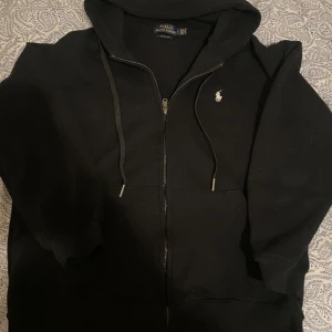 Svart hoodie från Polo Ralph Lauren - Det stor Storlek L fast den passar folk med storlek M så jag skulle säga att det är storlek M. Snygg svart hoodie från Polo Ralph Lauren med dragkedja och klassisk logga på bröstet. Perfekt för en avslappnad stil. Den har en justerbar huva och ribbade muddar för extra komfort.