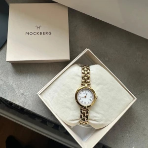 Mockberg klocka - Elegant guldfärgad klocka från Mockberg med en stilren vit urtavla. Klockan har en länkarmband i guldton och kommer i originalförpackning. Perfekt för att ge en touch av lyx till din stil.