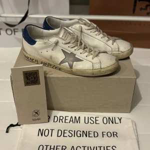 Golden Goose vita sneakers med blå detaljer - Snygga vita sneakers från Golden Goose med en ikonisk stjärna i silver på sidan och blå detaljer på hälen. Skorna har en vintage-look med snörning och en bekväm sula. Perfekta för en avslappnad stil.