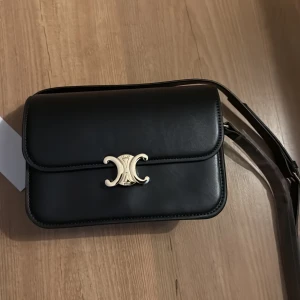 Svart axelväska med gulddetalj - Snygg svart axelväska med elegant guldfärgad detalj på framsidan. Väskan har en stilren design och justerbar axelrem. Perfekt för att ge en touch av lyx till din outfit.