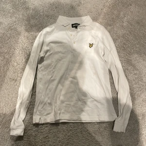 Vit långärmad pikétröja från Lyle & Scott - Snygg vit långärmad pikétröja från Lyle & Scott med klassisk krage och knappslå. Tröjan har en broderad logotyp på bröstet. 