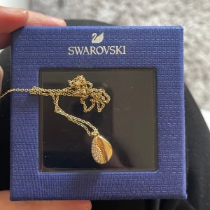 Guldhalsband med kristallhänge från Swarovski - Elegant guldhalsband från Swarovski med ett droppformat hänge dekorerat med gnistrande kristaller. Perfekt för att lägga till en touch av glamour till din look. Kedjan är fin och smidig, vilket gör den bekväm att bära hela dagen.