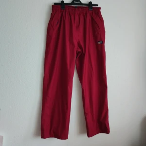 Röda täckbyxor från Rukka - Snygga röda täckbyxor från Rukka, perfekta för kyliga dagar. Byxorna har elastisk midja och justerbara benslut för optimal passform. Märkeslogga på sidan ger en sportig touch.
