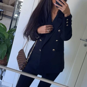 Svart kavaj med guldknappar - Snygg svart kavaj med dubbelknäppning och guldknappar. Perfekt för en stilren look. Kavajen har långa ärmar och en klassisk krage.