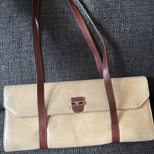 Beige axelväska från Armani - Elegant vintage beige axelväska från Armani med bruna läderremmar och ett stilrent metallspänne. Väskan har en strukturerad yta och ett rymligt inre med märkets logotyp på insidan. Värd 12.000