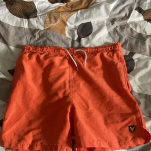 Orange badshorts från Lyle & Scott - Snygga orange shorts från Lyle & Scott med snörning i midjan. Perfekta för sommarens äventyr. De har en liten broderad logga på benet och är designade för juniorer i åldern 14/15 år.