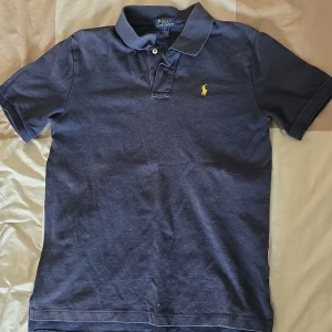 Mörkblå pikétröja från Ralph Lauren - Säljer en klassisk mörkblå pikétröja från Ralph Lauren med den ikoniska broderade loggan i gult på bröstet. Tröjan har korta ärmar och en stilren krage med knappar. Perfekt för en avslappnad men stilren look.