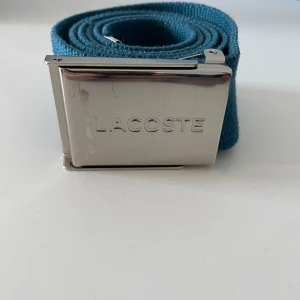 Blått bälte från Lacoste, 100cm - Snyggt blått bälte från Lacoste med en stilren silverfärgad spänne. Bältet har den klassiska Lacoste-loggan med krokodilen. Perfekt för att ge en färgklick till din outfit.