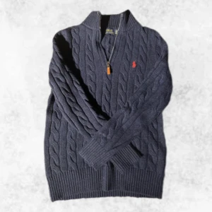 Kabelstickad halfzip från Ralph Lauren - Säljer en snygg mörkblå kabelstickad tröja från Ralph Lauren med dragkedja och klassiskt kabelstickat mönster. Tröjan har en liten röd broderad logga på bröstet och ribbade muddar. Perfekt för kyliga dagar! Den är i nyskick, endast testad. Vid några frågor eller funderingar är det bara att höra av sig.