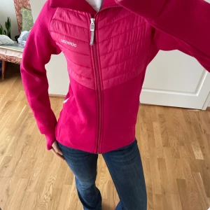 Rosa jacka från Didriksons - Snygg rosa jacka från Didriksons med dragkedja framtill. Jackan har en quiltad design på bröstet och långa ärmar. Perfekt för kyligare dagar med sin varma och bekväma passform. Storlek 170 men passar en XS!!