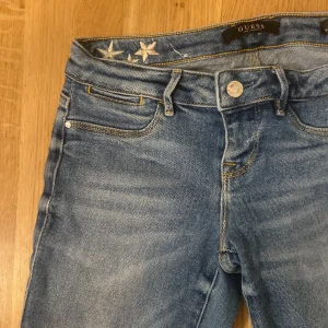 Blå jeans med stjärndetaljer från Guess - Snygga blå jeans från Guess med unika stjärndetaljer broderade vid midjan. Klassisk femficksdesign med en knapp och dragkedja framtill. Perfekta för en trendig look. Sånna typer av jeans är väldigt populära och trendiga just nu. De är lite tajta vid benen därför kan man sy ut de så att de blir mer utsvängda om man vill. Säljer dessa eftersom har aldrig använt de pågrund av att de är för små. Detta är storlek 25 i vuxen storlek då det är nog xs/s är rätt osäker. 