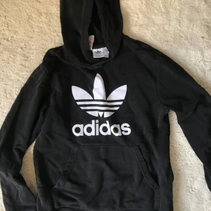 Svart hoodie från Adidas - Säljer en klassisk svart hoodie från Adidas med den ikoniska loggan i vitt på bröstet. Tröjan har en bekväm huva och en praktisk magficka. Perfekt för en avslappnad stil.