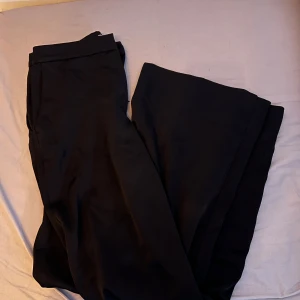 Svarta kostymbyxor - Snygga svarta kostymbyxor i storlek S från H&M i glansigt material. Byxorna har en klassisk design med raka ben och knappar i midjan.