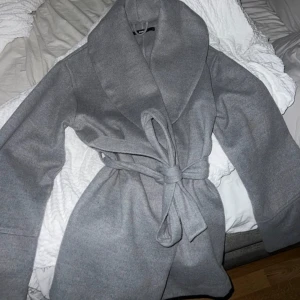 Grå kappa från BikBok - Säljer en stilren grå kappa från BikBok i storlek S. Den har en elegant knytning i midjan och en bred krage som ger en sofistikerad look. Perfekt för kyligare dagar. Använd en gång 