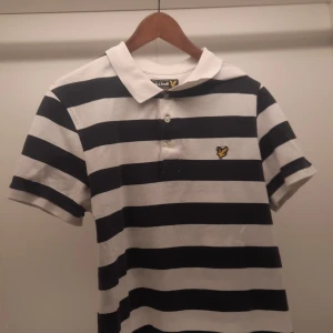 Randig pikétröja från Lyle & Scott - Säljer en stilren randig pikétröja från Lyle & Scott i svart och vit. Tröjan har korta ärmar och en klassisk krage med knappar. Den ikoniska loggan är broderad på bröstet. Perfekt för en avslappnad och snygg look.