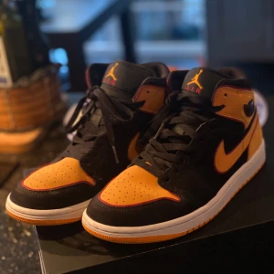 Nike Air Jordans i svart och orange - Snygga Nike Air Jordans 1 mid i en cool kombination av svart och orange. Skorna har en klassisk hög design med snörning och den ikoniska Air Jordan-loggan på sidan. Perfekta för att ge din stil ett lyft. Mycket bra skick på skorna och är knappt använda. 