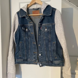 Jeansjacka från Levi's - Snygg jeansjacka från Levi's med gråa ärmar och huva i sweatshirtmaterial. Jackan har knappar framtill och två bröstfickor med knapp. Perfekt till våren. 