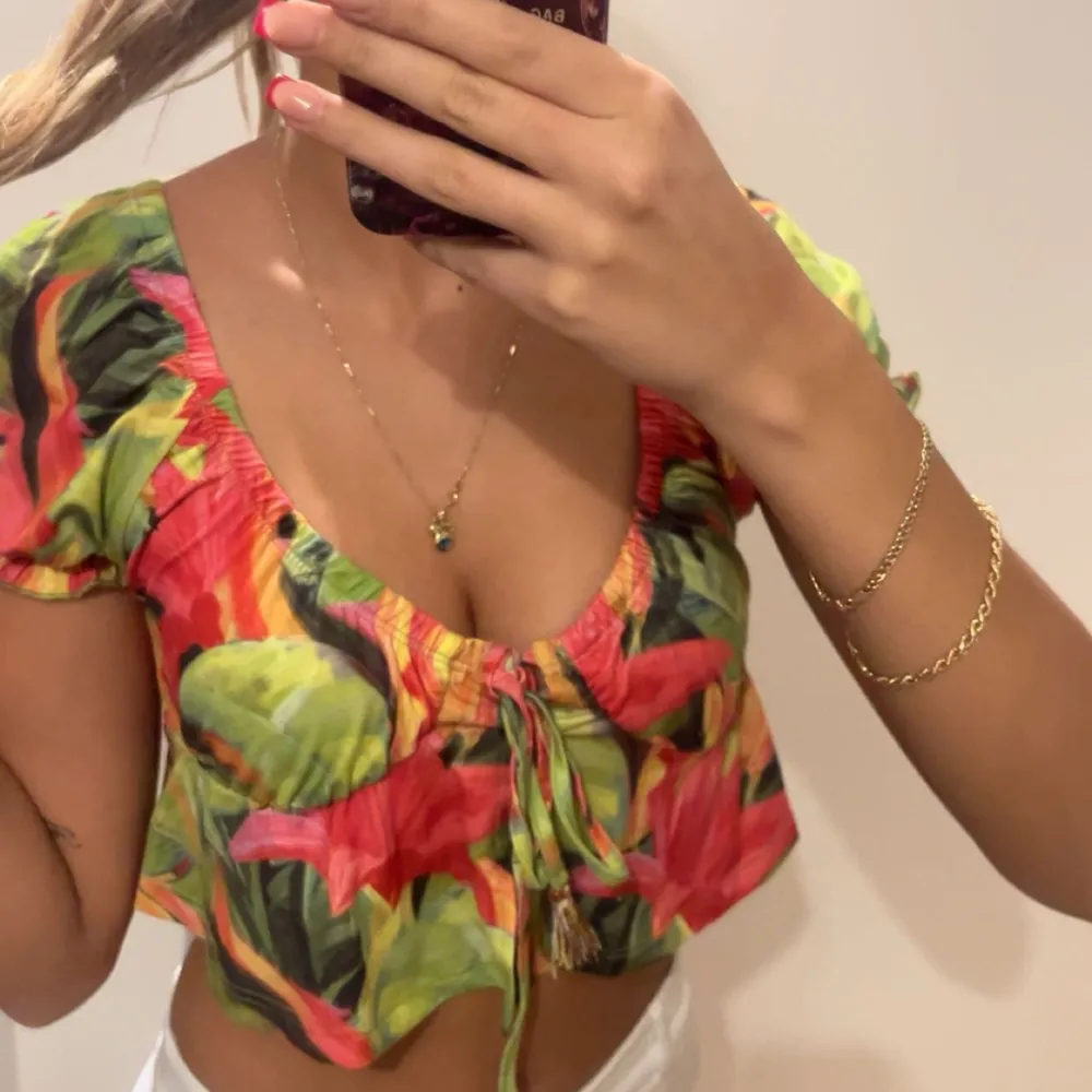 Säljer en färgglad blommig croppad topp med korta ärmar och knytning framtill. Perfekt för sommardagar med sitt livfulla mönster i grönt, rött och svart. Toppen har en v-ringad design som ger en feminin touch.. Yläosat.